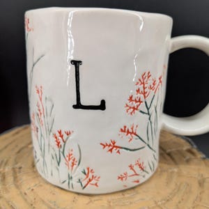 May include: White ceramic mug with a black letter "L" and floral design in red and green. The mug has a curved handle and is set on a wooden surface. The mug is a unique and personalized gift.