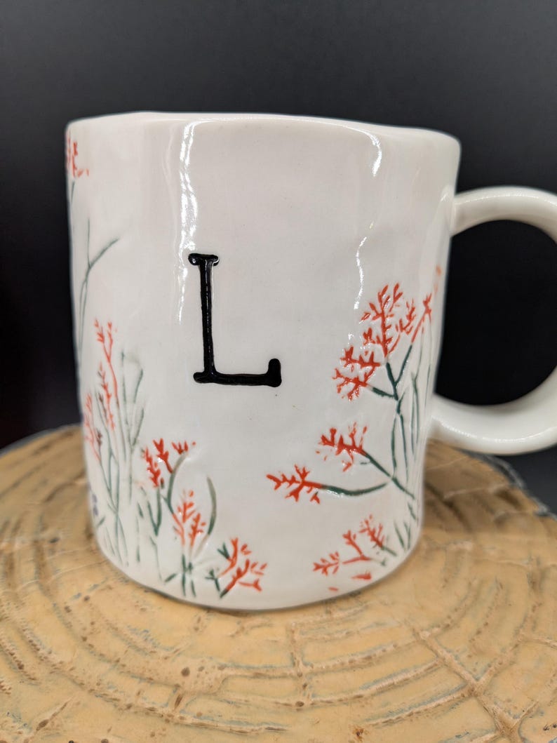 May include: White ceramic mug with a black letter "L" and floral design in red and green. The mug has a curved handle and is set on a wooden surface. The mug is a unique and personalized gift.