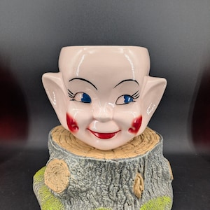 May include: A vintage ceramic planter shaped like a smiling face with blue eyes, red cheeks, and a red mouth. The planter sits atop a tree stump base with green moss and wood grain details. A unique decorative item.