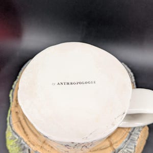 May include: White ceramic mug with a handle, featuring the text "by ANTHROPOLOGIE" on the bottom. The mug is placed on a decorative, tree-stump-shaped coaster with a light brown and green color scheme.