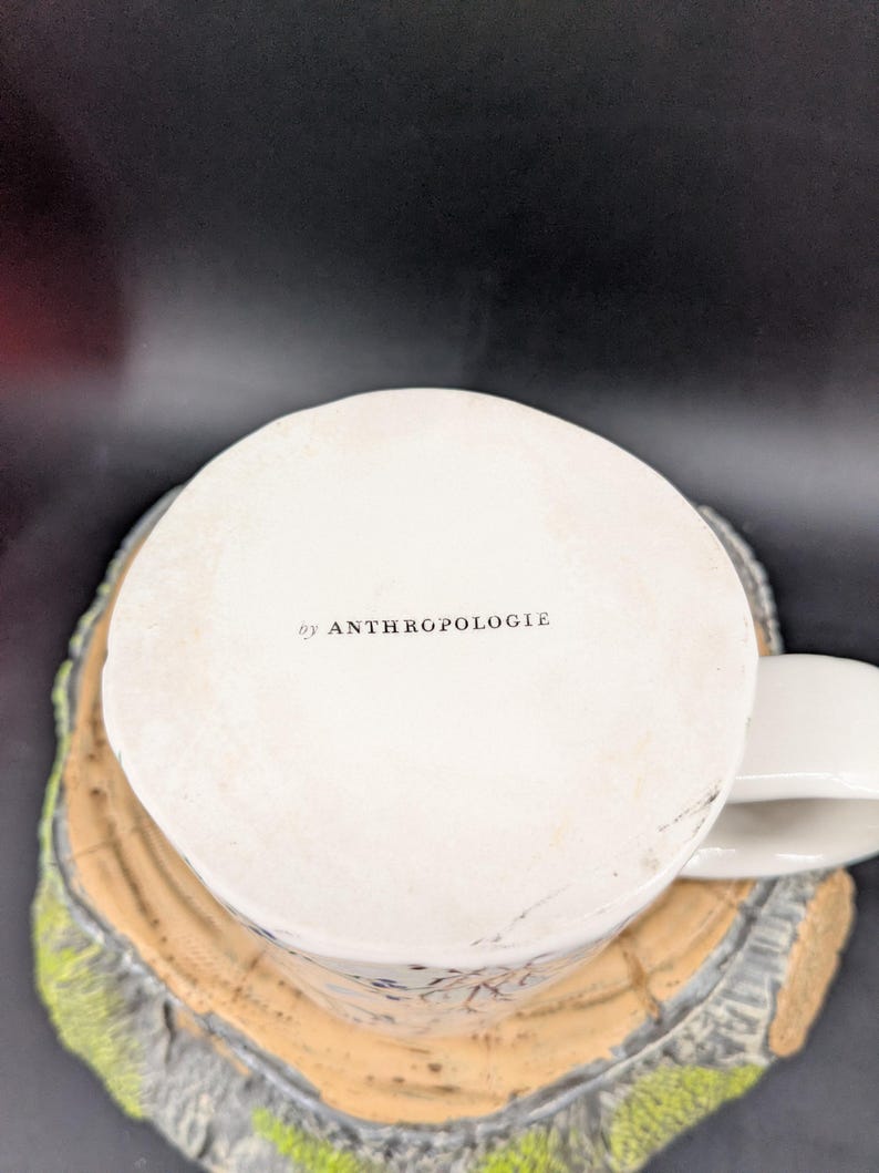 May include: White ceramic mug with a handle, featuring the text "by ANTHROPOLOGIE" on the bottom. The mug is placed on a decorative, tree-stump-shaped coaster with a light brown and green color scheme.