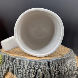 May include: A white ceramic mug with a handle, viewed from above. The mug is set on a decorative tree stump base with a textured, gray and brown finish. The interior of the mug is smooth and white.