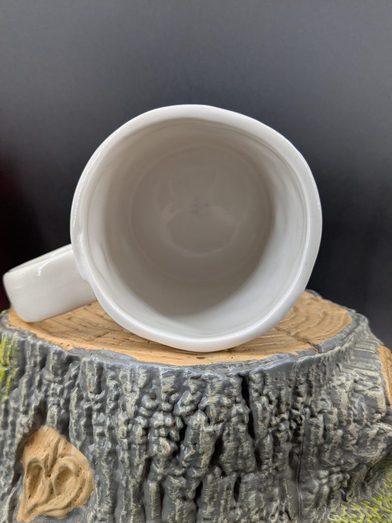 May include: A white ceramic mug with a handle, viewed from above. The mug is set on a decorative tree stump base with a textured, gray and brown finish. The interior of the mug is smooth and white.