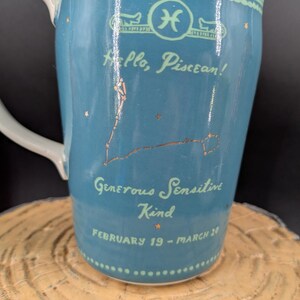 K&ouml;nnte beinhalten: Eine t&uuml;rkisfarbene Keramiktasse mit wei&szlig;em Henkel, die das Sternzeichen Fische und die Konstellation in Gold zeigt. Die Tasse tr&auml;gt den Text "Hello, Piscean!" und "Generous, Sensitive, Kind, February 19 - March 20".