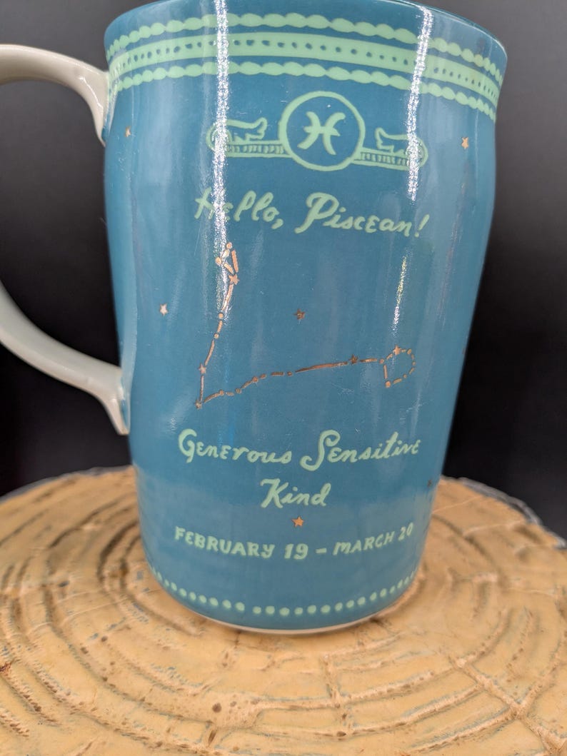 K&ouml;nnte beinhalten: Eine t&uuml;rkisfarbene Keramiktasse mit wei&szlig;em Henkel, die das Sternzeichen Fische und die Konstellation in Gold zeigt. Die Tasse tr&auml;gt den Text "Hello, Piscean!" und "Generous, Sensitive, Kind, February 19 - March 20".