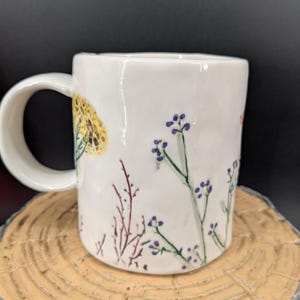 May include: White ceramic mug with a curved handle, decorated with hand-painted floral designs in yellow, purple, and red. The mug is set on a wooden surface against a dark background. The mug is approximately 4 inches tall.