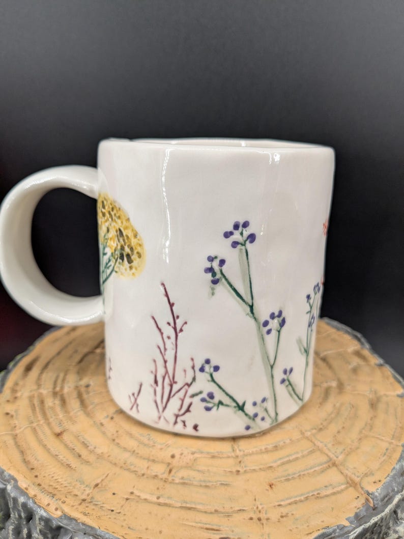 May include: White ceramic mug with a curved handle, decorated with hand-painted floral designs in yellow, purple, and red. The mug is set on a wooden surface against a dark background. The mug is approximately 4 inches tall.