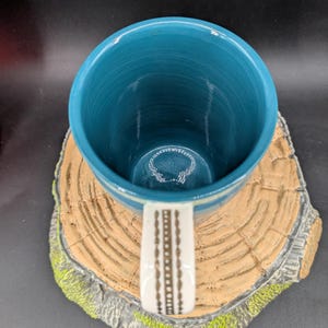K&ouml;nnte beinhalten: Eine t&uuml;rkisfarbene Keramiktasse mit einem wei&szlig;en Griff mit einem braunen geometrischen Muster. Das Innere der Tasse ist ein leuchtendes T&uuml;rkis, und sie steht auf einem dekorativen Untersetzer in Form eines Baumstumpfes mit gr&uuml;nen Moosakzenten.