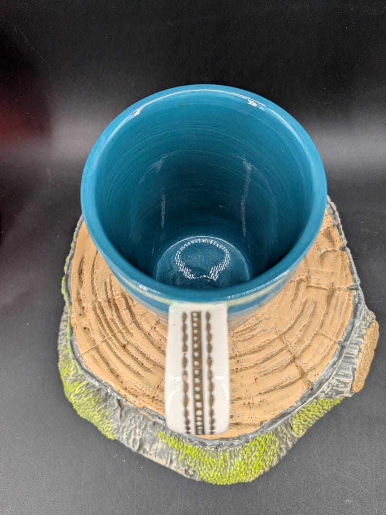 K&ouml;nnte beinhalten: Eine t&uuml;rkisfarbene Keramiktasse mit einem wei&szlig;en Griff mit einem braunen geometrischen Muster. Das Innere der Tasse ist ein leuchtendes T&uuml;rkis, und sie steht auf einem dekorativen Untersetzer in Form eines Baumstumpfes mit gr&uuml;nen Moosakzenten.