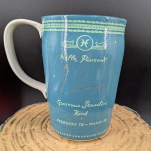 K&ouml;nnte beinhalten: Eine blaue Keramiktasse mit wei&szlig;em Henkel, die das Sternzeichen Fische und die Konstellation zeigt. Die Tasse tr&auml;gt den Text "Hello, Piscean!" und "Generous, Sensitive, Kind, February 19 - March 20."