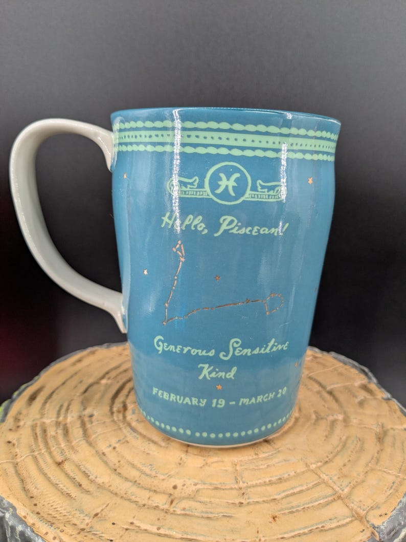 K&ouml;nnte beinhalten: Eine blaue Keramiktasse mit wei&szlig;em Henkel, die das Sternzeichen Fische und die Konstellation zeigt. Die Tasse tr&auml;gt den Text "Hello, Piscean!" und "Generous, Sensitive, Kind, February 19 - March 20."