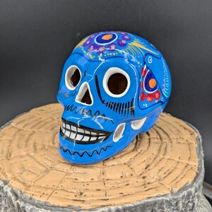 May include: A blue ceramic skull with colorful painted details. The skull is decorated with flowers and swirls. It is a traditional Mexican Day of the Dead decoration.
