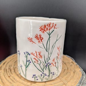 May include: White ceramic cup with a hand-painted floral design. The cup features red, green, and purple flowers and foliage. The cup is cylindrical and sits on a wooden surface.