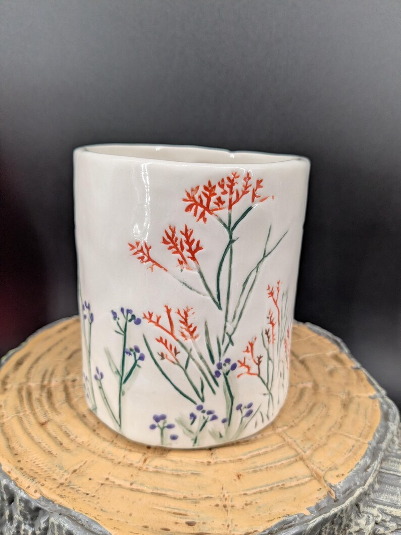 May include: White ceramic cup with a hand-painted floral design. The cup features red, green, and purple flowers and foliage. The cup is cylindrical and sits on a wooden surface.