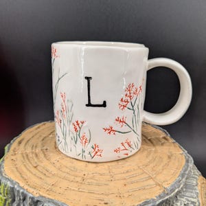 May include: White ceramic mug with a black letter "L" and floral design. The mug features red and green floral accents. The mug is sitting on a wooden surface.