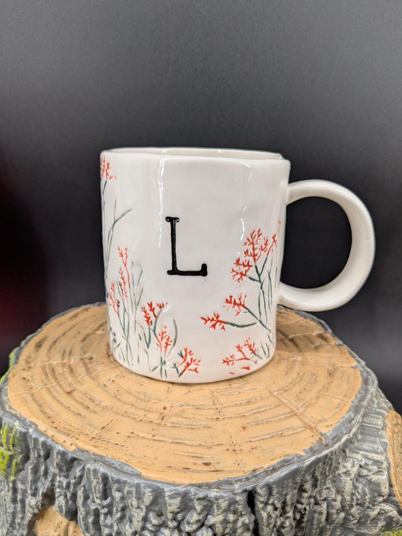 May include: White ceramic mug with a black letter "L" and floral design. The mug features red and green floral accents. The mug is sitting on a wooden surface.