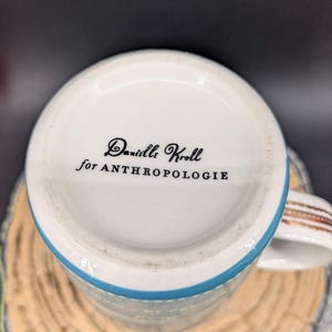 K&ouml;nnte beinhalten: Wei&szlig;er Keramikbecher mit blauem Rand und dekorativem Henkel. Auf der Unterseite des Bechers steht in schwarzer Schrift "Danielle Kroll for ANTHROPOLOGIE". Der Becher steht auf einer Holzoberfl&auml;che.