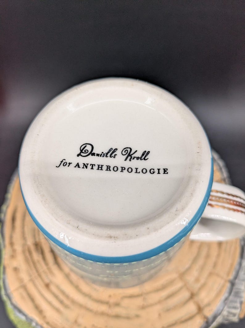 K&ouml;nnte beinhalten: Wei&szlig;er Keramikbecher mit blauem Rand und dekorativem Henkel. Auf der Unterseite des Bechers steht in schwarzer Schrift "Danielle Kroll for ANTHROPOLOGIE". Der Becher steht auf einer Holzoberfl&auml;che.