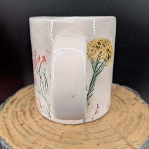 May include: White ceramic mug with a rectangular handle, decorated with hand-painted floral designs in red, yellow, and green. The mug is set on a wooden surface, showcasing the detailed artwork and craftsmanship.