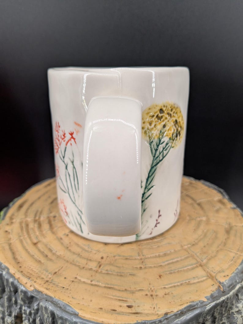 May include: White ceramic mug with a rectangular handle, decorated with hand-painted floral designs in red, yellow, and green. The mug is set on a wooden surface, showcasing the detailed artwork and craftsmanship.