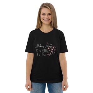 Peut inclure: Un t-shirt noir à manches courtes avec le texte blanc "Making Magic One Stitch at a Time" accompagné d'un motif rose représentant une aiguille et du fil formant une boucle, agrémenté de petites étoiles roses à quatre branches éparpillées.