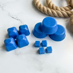 May include: A set of blue wax melts in various shapes, including diamonds and circles. The wax melts are arranged on a white marble surface.