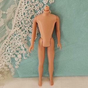 May include: A tan-colored doll body with articulated arms and legs. The doll's head is missing. The doll is positioned in front of a light blue background and a white lace trim.