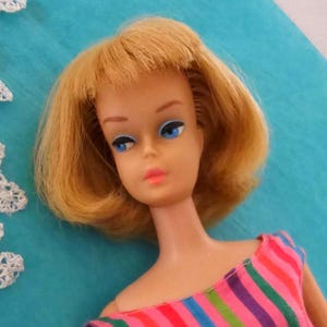 May include: A vintage doll with blonde hair and blue eyes. The doll is wearing a striped top with pink, purple, green, and blue stripes. The doll's face has a classic, retro look, with painted features.