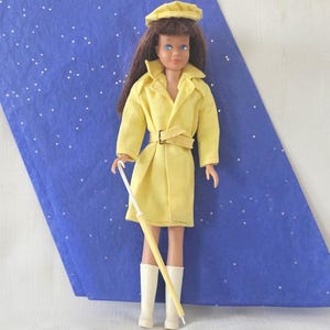 May include: A vintage doll wearing a yellow raincoat, matching hat, and white boots. The doll has brown hair and blue eyes, holding a yellow and white umbrella. The doll is posed against a blue background with white dots.