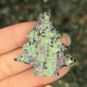 May include: A carved gemstone Christmas tree figurine. The tree is green with black and purple speckles. The figurine is held in a hand.