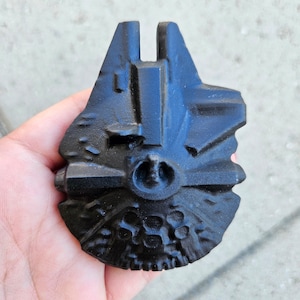 May include: A black metal model of the Millennium Falcon spaceship from Star Wars. The model is detailed with the iconic features of the ship, including the cockpit, engines, and wings.