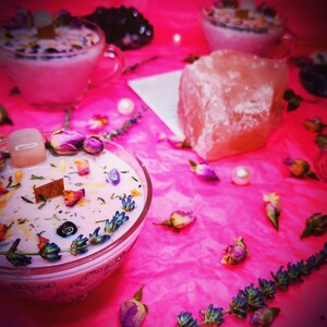 May include: Two pink glass candle jars with floral and crystal accents on a pink surface. The candles are surrounded by dried flowers and crystals.