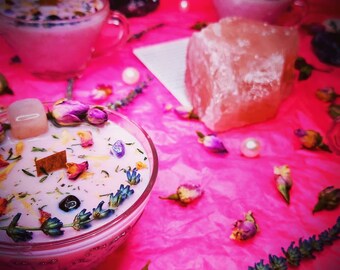May include: Two pink glass candle jars with floral and crystal accents on a pink surface. The candles are surrounded by dried flowers and crystals.