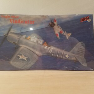 May include: A model kit of a Vought SB2U Vindicator aircraft, a US Navy dive bomber, with a red, white, and blue color scheme. The box art features the aircraft in flight with a Japanese fighter plane in the background. The box is labeled "Vought SB2U Vindicator" and "HiPM".