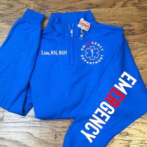May include: A royal blue quarter-zip sweatshirt with "EMERGENCY" printed in white and red on the sleeve. The front features the text "Lisa, RN, BSN" and an emergency medical services emblem.
