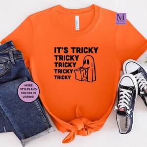 May include: Orange t-shirt with black text that reads "It's Tricky Tricky Tricky Tricky Tricky". A black and white illustration of a ghost holding a bag of candy is next to the text.