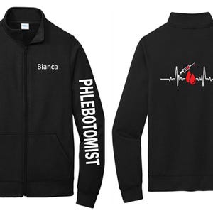 May include: Black zippered jacket with a white heartbeat graphic on the back and the words "PHLEBOTOMIST" on the left sleeve. The name "Bianca" is embroidered on the front of the jacket.