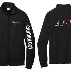 Può includere: Giacca nera con zip con testo bianco "Cardiologia" sulla manica e "Bianca" sul petto. Il retro presenta un cuore rosso e una linea del battito cardiaco con la scritta "The Beat Goes On."