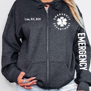 May include: Dark grey zip-up hoodie with white text. The left chest reads "Lisa, RN, BSN." The right sleeve displays "EMERGENCY" vertically. The front features "EMERGENCY DEPARTMENT" around a medical symbol. A casual, comfortable garment for medical professionals.