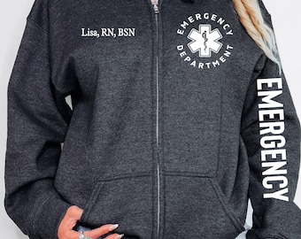 May include: Dark gray zip-up hoodie with white text. The left chest reads "Lisa, RN, BSN." The right sleeve displays "EMERGENCY" vertically. The front features "EMERGENCY DEPARTMENT" around a medical symbol. A casual, comfortable garment for medical professionals.