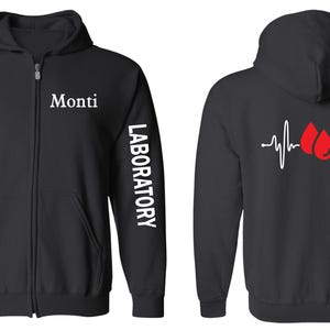 Puede incluir: Sudadera negra con cremallera y capucha, con la palabra "Monti" en la parte delantera y la palabra "LABORATORY" en la manga izquierda. La parte posterior de la sudadera tiene un gráfico de latido del corazón blanco con dos gotas de sangre rojas.