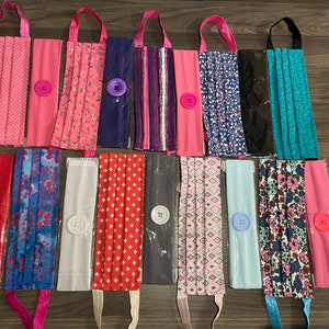 May include: A collection of 12 face masks with various patterns and colours, including pink, red, blue, and green. Each mask has a button sewn on the side and is attached to a headband.