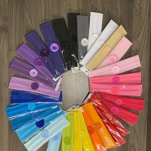 May include: A selection of colourful fabric headbands with buttons. The headbands are arranged in a circle, with the buttons facing outwards. The headbands are made from a soft, stretchy fabric and are ideal for keeping hair out of your face.