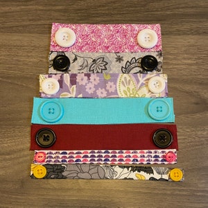 May include: A set of six fabric face mask extenders with different patterns and colours. The extenders have buttons on each end for attaching to a face mask.