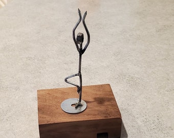 May include: A metal sculpture of a person in a yoga pose, standing on a wooden base. The sculpture is made of wire and metal.