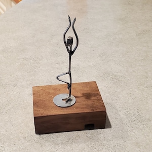 May include: A metal sculpture of a person in a yoga pose, standing on a wooden base. The sculpture is made of wire and metal.