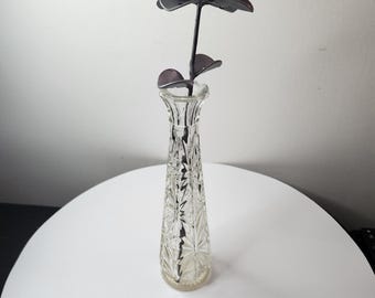 May include: A dark gray metal flower sculpture with rounded petals and two leaves on a thin stem, placed inside a tall, clear glass bud vase featuring a vertical geometric cut pattern and a flared rim, standing upright on a smooth surface.