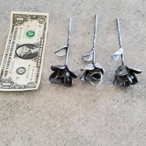 May include: Three metal roses, each with a different size and shape, are arranged in a row on a gray surface. A dollar bill is placed below the roses for scale.