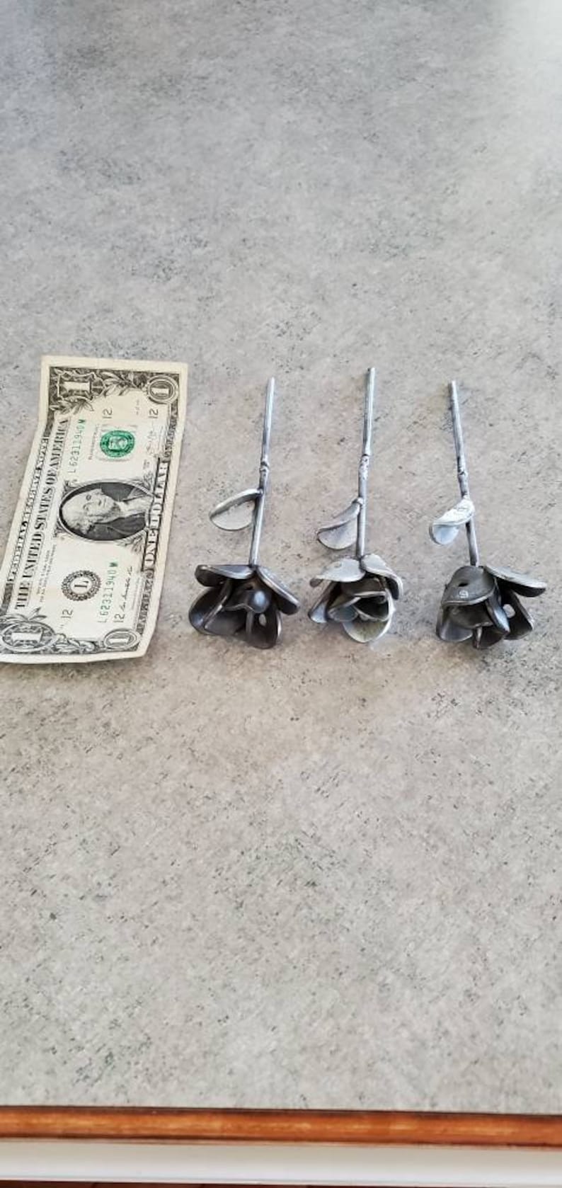 May include: Three metal roses, each with a different size and shape, are arranged in a row on a gray surface. A dollar bill is placed below the roses for scale.