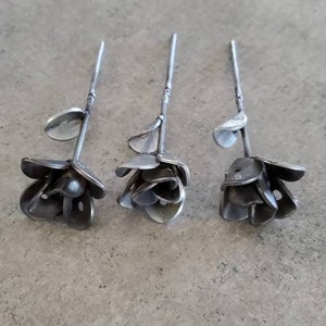 May include: Three metal roses with stems, each crafted from recycled metal, are arranged on a gray surface. The roses are a unique and artistic way to add a touch of nature to any home.